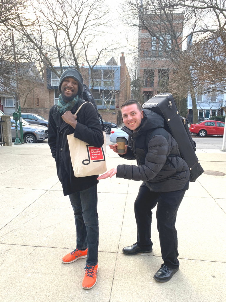 Two male artist teachers outside. One is carrying a CSMA branded bag and the other is carrying an instrument case and motioning to the CSMA bag.